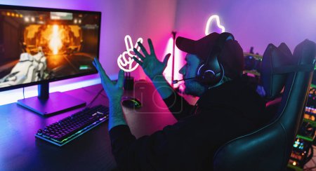 agresivo y gritando Gamer Jugar en primera persona Shooter Juego de vídeo en línea en su potente ordenador personal. La habitación y la PC tienen luces led de neón coloridas. Joven lleva una gorra en casa