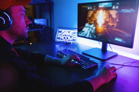 Photo for Shot of the Professional Gamer Playing in First-Person Shooter Online Video Game on His Personal Computer. Room Lit by Neon Lights in Retro Arcade Style. Online Cyber e-Sport Internet Championship - Royalty Free Image