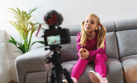 Téléchargez les photos : Belle jeune fille en tenue décontractée rose souriant à la caméra et parlant sur la prise de vue vidéo avec la technologie. Enfants Influence des médias sociaux ou concept jeune créateur de contenu dans un style décontracté relax à la maison. - en image libre de droit