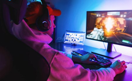 Photo for Professional Gamer Playing First-Person Shooter Online Video Game on His Powerful Personal Computer. Room and PC have Colorful Neon Led Lights. Young Man is Wearing a Cap at Home. - Royalty Free Image