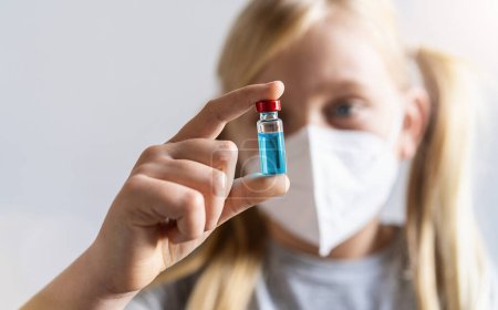 Photo for Blonde girl with face mask shows a Vaccine bottle with blue poison for Covid-19 vaccine at a vaccination center. - Royalty Free Image