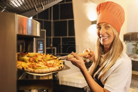 Photo for Traditional female pizza baker holding a fresh pizza from the stone oven on peel at italian pizzeria - Royalty Free Image
