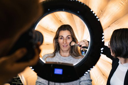 Téléchargez les photos : Maquilleur poudres un visage de modèles posant à la caméra à travers une lampe à lumière annulaire à une séance de photo avec lumière annulaire - en image libre de droit