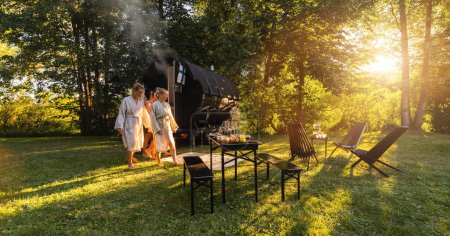 Entspannte Menschen in Bademänteln kommen aus einer hölzernen Fasssaunahütte in einem üppig grünen Park bei Sonnenuntergang. Sie sind entspannt, lachen und genießen den Urlaub beim Relaxen in der finnischen Saunakabine.