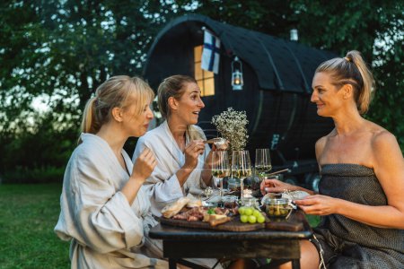 Freunde in Bademänteln genießen eine Mahlzeit und Wein in der Nähe eines finnischen Saunafässers im Freien