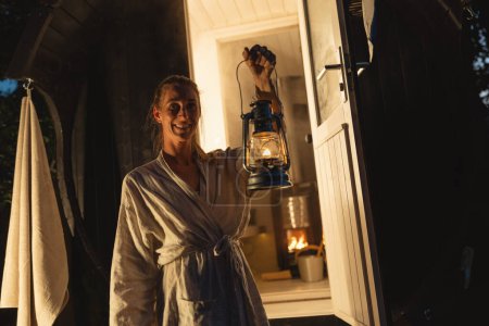 Mujer sonriente sosteniendo una linterna en la entrada de una sauna de barril finlandesa durante la noche.