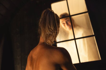 femme à l'extérieur d'un baril finlandais sauna fenêtre sourit à son amie à l'intérieur, vapeur visible.