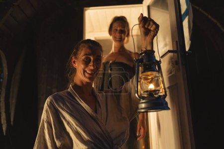 Téléchargez les photos : Sauna finlandais baril avec porte ouverte, femme à l'intérieur souriant à un ami tenant une lanterne. - en image libre de droit