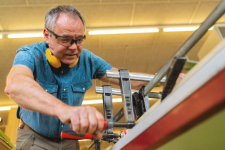 Carpintero enfocado con protección auditiva trabajando con abrazaderas y madera en un taller