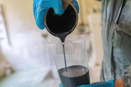 Foto de Primer plano de las manos vertiendo pintura negra en una taza de medir transparente con guantes azules - Imagen libre de derechos