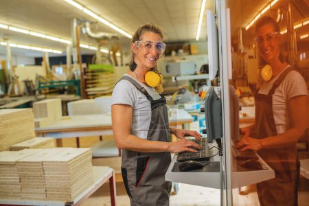 Mujer sonriente con orejeras usando una computadora en un taller de carpintería