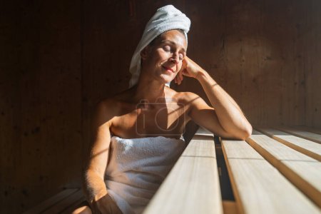 Frau in finnischer Sauna lehnt sich zurück, die Augen geschlossen, mit einem Handtuch um den Kopf gewickelt, sonnenbeschienen in einem Kurort