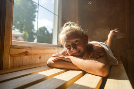 Lächelndes Kind auf einer Saunabank liegend, Sonnenlicht strömt aus einem Fenster herein