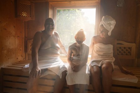 Famille dans un sauna finlandais riant ensemble, homme à gauche, femme avec serviette sur la tête à droite, enfant au milieu à l'hôtel wellness