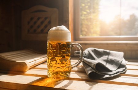 Bierkrug mit schaumigem Bier ruhen auf einem Felsvorsprung in der Sauna und fangen das Sonnenlicht ein. In der Nähe finnische Saunahüte auf einer Holzbank. Kur- und Wellness-Konzept Image