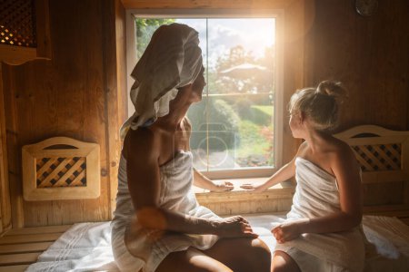 Back view of a mother and daughter in a finnish sauna, looking out a window, wrapped in towels at a spa wellness hotel