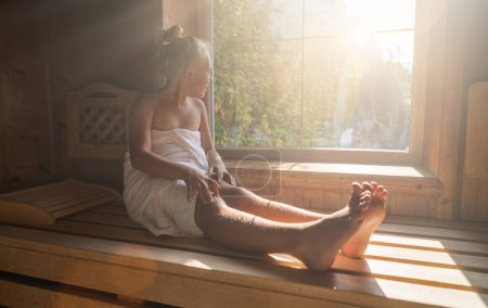 Niño sentado en un banco de sauna finlandés, envuelto en una toalla, mirando por una ventana soleada a un hotel spa