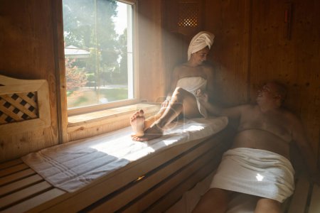 Couple relaxing in a sauna, woman with a towel on her head, both seated and smiling at spa hotel