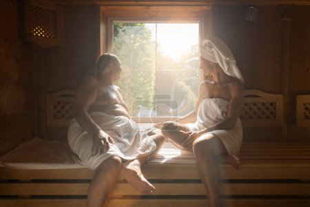 Hombre y mujer sentados uno frente al otro en una sauna de acabado, ambos envueltos en toallas, ventana soleada en el hotel spa