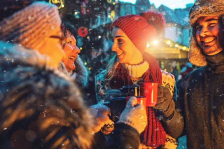 Des amis joyeux dégustant du vin chaud et du chocolat chaud dans un marché de Noël enneigé