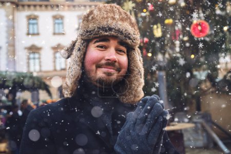 Foto de Hombre con sombrero de piel y abrigo de invierno sonriendo suavemente, copos de nieve cayendo a su alrededor, luces del mercado de Navidad en el fondo. - Imagen libre de derechos