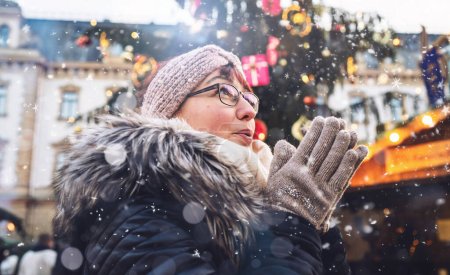 Foto de Mujer con gafas y un sombrero de invierno aplaude las manos para calentar, caída de nieve, mercado de Navidad - Imagen libre de derechos