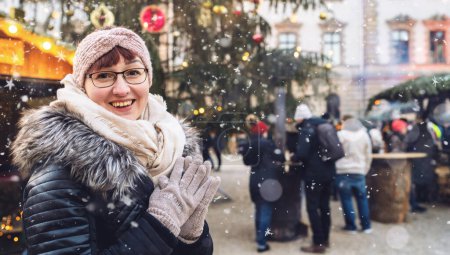 Foto de Mujer joven disfrutando de un mercado navideño nevado, vistiendo ropa de invierno y gorro, sonriendo en cámara fotográfica con espacio para su texto individual. - Imagen libre de derechos