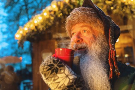 Foto de Hombre sonriente con una barba sosteniendo una taza roja de vino caliente o chocolate caliente en un mercado de Navidad, luces festivas en el fondo - Imagen libre de derechos