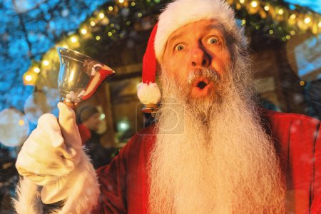 Foto de Santa Claus con mirada sorprendida, tocando una campana en un mercado de Navidad, luces festivas telón de fondo - Imagen libre de derechos