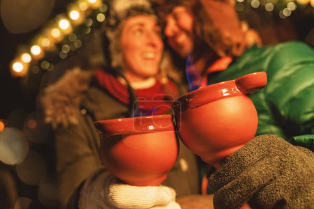 Foto de Riendo pareja disfrutando de pinzas de fuego puñetazo en un mercado de Navidad, sosteniendo tazas rojas humeantes con llamas azules. - Imagen libre de derechos