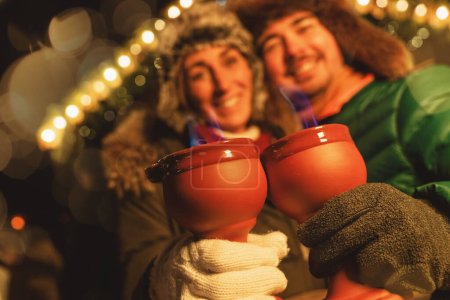 Foto de Pareja sonriente en un mercado de Navidad, sosteniendo tazas rojas con llamas azules de pinzas de fuego ponche, luces festivas detrás. - Imagen libre de derechos