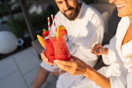 Couple in white robes toasting with fruit garnished cocktails in sunlight at spa hotel