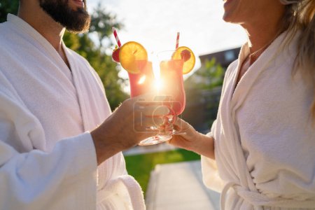 Couple in white bathrobes clinking red cocktails with a sunset behind at spa hotel