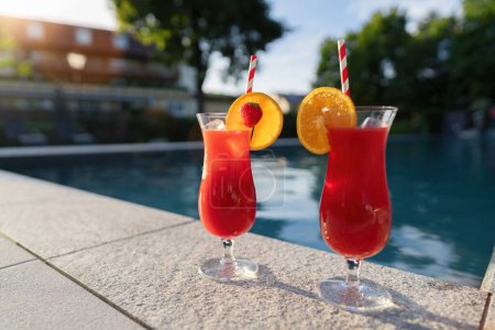 Two red cocktails with fruit garnishes by a poolside, reflecting the warm sunset light