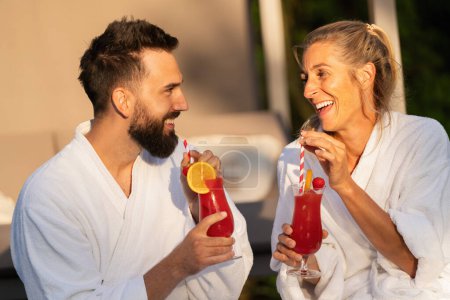 Two people in white robes laughing and drinking cocktails in the sunlight at a wellness hotel
