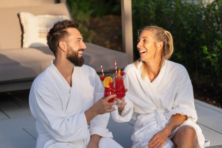 joyful couple in white bathrobes clinking glasses with red drinks, outdoors at golden hour at a hotel