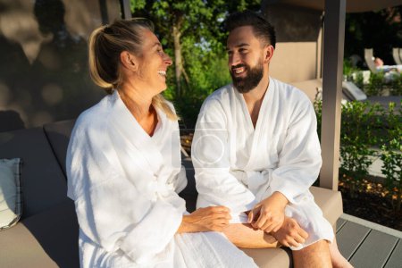 Lächelndes Paar in weißen Bademänteln genießt einen sonnigen Tag im Freien auf der Liege eines Paares in einem Wellnesshotel