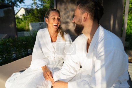 Zwei Menschen in weißen Bademänteln sitzen auf der Liege eines Paares im Freien und lachen gemeinsam in einem Hotel