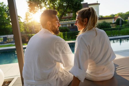 Foto de Pareja enamorada mirándose el uno al otro albornoces blancos disfrutando de la puesta de sol en una piscina en un hotel spa wellness - Imagen libre de derechos