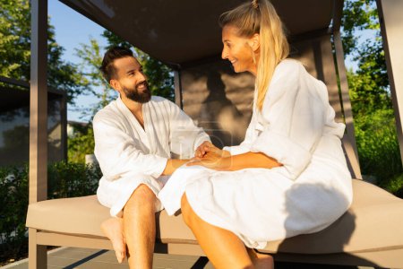 Foto de Pareja sonriente enamorada en batas blancas sentada en una tumbona al aire libre a la luz del sol en el hotel spa wellness - Imagen libre de derechos