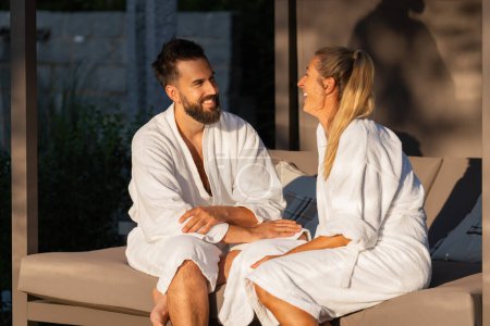 glückliches Paar in weißen Bademänteln auf einer Liege im Freien im Sonnenlicht im Wellnesshotel