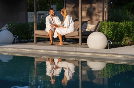 Paar in Bademänteln auf einer Liege am Pool, Spiegelung im Wasser, Outdoor-Kulisse in einem Wellness-Resort 