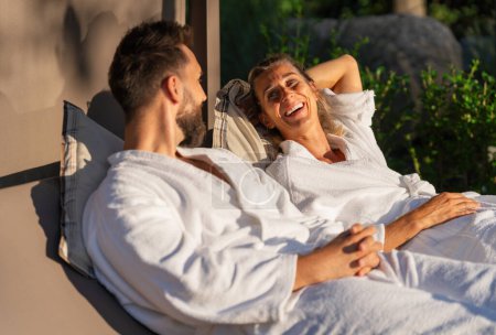 Paar in weißen Bademänteln entspannt und lacht gemeinsam auf Liegen im Sonnenlicht in einem Wellness-Hotel