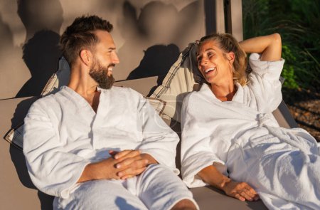Paar in weißen Bademänteln im Liegen auf einer Liege im Sonnenlicht eines Wellnesshotels 