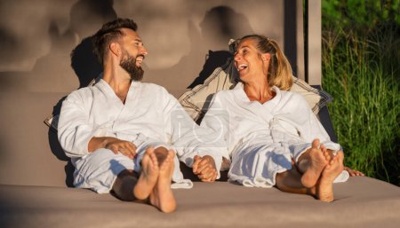 Paar in weißen Bademänteln im Liegen auf einer Liege im Sonnenlicht eines Wellnesshotels 