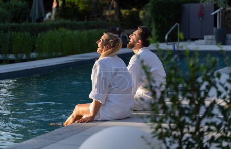 Paar in weißen Bademänteln sitzt am Pool und blickt auf und lacht einen Tag in einem Wellnesshotel