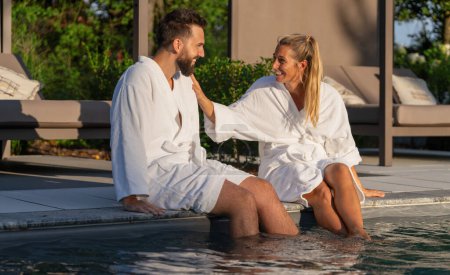 Paar in weißen Bademänteln sitzt mit Füßen im Pool und genießt einen sonnigen Tag im Wellnesshotel