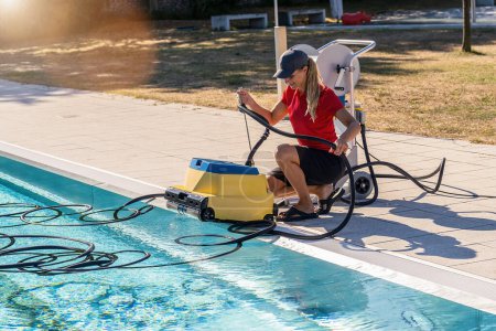 técnico de la piscina agachado junto a una piscina, el manejo de una manguera limpiador de piscina robótica