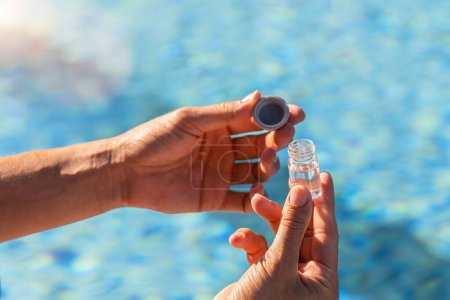 Hands of a pool technician holding a vial of water with an open cap for pH testing, over a swimming pool background