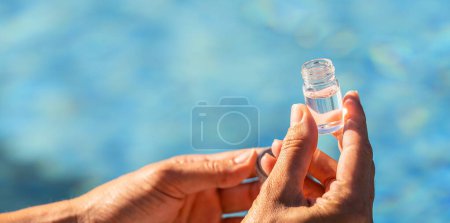 Hands of a pool technician holding a small open vial with clear water sample for pH testing against a pool background